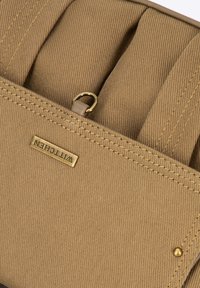 Borsa in tela marrone con hardware dorato, caratterizzata da un'etichetta con logo in evidenza, corpo strutturato e dettagli di cucitura visibili.
