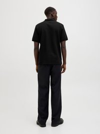 Mężczyzna stoi tyłem, ubrany w czarną koszulę polo z krótkim rękawem, czarne spodnie i czarne buty na tle jasnego, jednolitego tła.