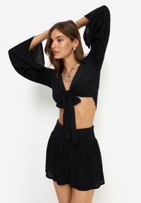 Μαύρο τοπ καλλιέργειας με μακριά, καμπανωτά μανίκια και λεπτομέρεια με δέσιμο μπροστά. Συνδυασμένο με μαύρη, ραμμένη μίνι φούστα με ψηλή μέση. Λεία υφή.