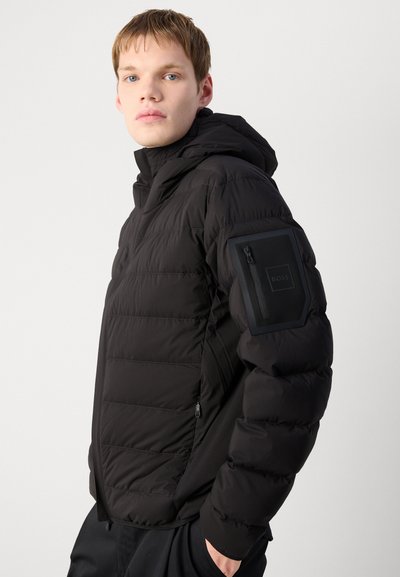 Doudoune noire avec un matelassage horizontal, une capuche, et une poche zippée sur le bras gauche arborant un logo. Tissu doux et texturé.