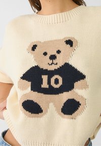 Pull couleur crème avec un motif de nounours tricoté portant un t-shirt noir affichant le numéro "10" et des accents contrastants couleur brun clair.