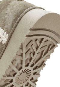 Suela de zapato con un intrincado patrón de tracción geométrico en beige claro. El material parece texturizado y similar al caucho, con acentos de suave gamuza alrededor.