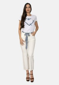 Camiseta blanca de manga corta con diseño de corazón azul. Pantalones color crema con cinturón de lazo de patrón azul. Sandalias de tacón marrones.