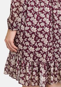 Floral Kleid in tiefem Burgunderrot mit weißem Blumenmuster. Durchscheinender Stoff mit einem Rüschensaum und Knopfmanschetten an den Ärmeln.