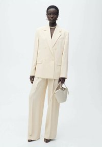 Sacou bej cu două rânduri de nasturi, purtat cu pantalonii evazați. Material fin, croială ajustată și design minimalist.