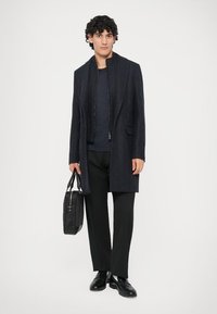 Abrigo gris oscuro con cuello alto y bolsillos delanteros, combinado con una camiseta negra, pantalones negros y zapatos negros. Se lleva una bolsa negra en la mano.