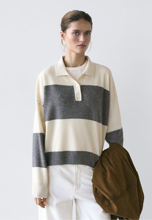 Junge Frau mit zurückgebundenem Haar, die einen cremefarbenen und grauen gestreiften Pullover und weiße Hosen trägt, hält eine braune Jacke in einer Hand.