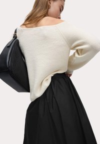 Pullover in maglia crema con design scollato sulle spalle, abbinato a una gonna nera. Una borsa a tracolla nera con texture è appoggiata su una spalla.