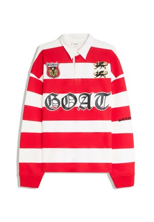 Junger Mann trägt ein rot-weiß gestreiftes Rugbyshirt mit dem Text "GOAT" und eine schwarze Kappe mit dem Text "AZURE", steht vor einem schlichten beigen Hintergrund.