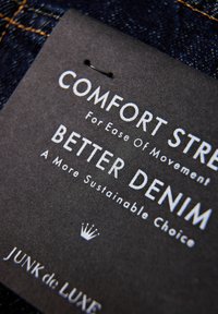 Czarny materiałowy metka z białym tekstem promującym "Komfortowy Stretch" i "Lepszy Dżins." Zawiera symbol korony oraz nazwę marki "JUNK de LUXE."