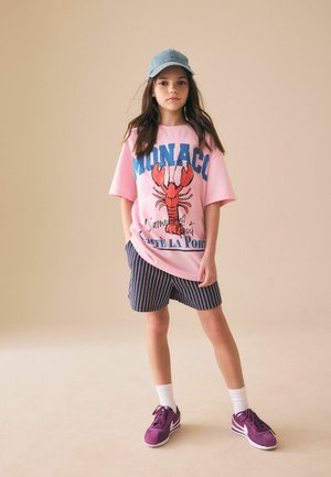 Noor tüdruk sinise mütsi, roosa suure suurusega Monaco särgi homaarigraafikuga, triibuliste lühikeste pükstega, valgete sokkidega ja lilla spordijalatsitega seismas beeži seina ääres.