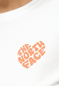 Πορτοκαλί κείμενο "The North Face" διατεταγμένο σε σχήμα καρδιάς πάνω σε λευκό ύφασμα, πιθανώς το μπροστινό μέρος ενός μπλουζακιού.