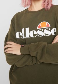 Olive green sweatshirt with white "ellesse" logo and orange accent. Features a crew neckline and ribbed cuffs. Soft, textured fabric.