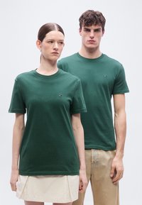 Γυναίκα και άνδρας στέκονται δίπλα-δίπλα, φορώντας ταιριαστά σκούρα πράσινα T-shirts με μικρά λογότυπα, η γυναίκα σε μπεζ φούστα και ο άνδρας σε ανοιχτό καφέ παντελόνι.