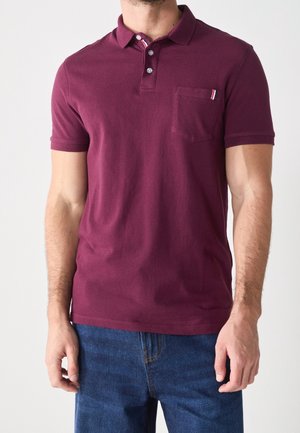 Mann trägt ein kurzärmeliges, kastanienbraunes Polohemd mit Brusttasche und blaue Jeans, steht vor einem schlichten hellen Hintergrund.