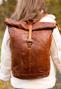 Zaino in pelle marrone con design a roll-top, dotato di una chiusura in metallo e texture liscia. La borsa presenta cuciture ordinate e uno stile minimalista.