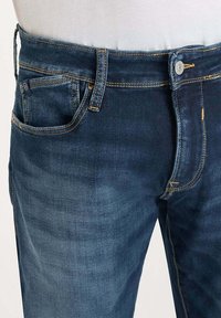 Jeans de mezclilla en un lavado azul oscuro con costuras amarillas en contraste, cierre de botón y diseño de cinco bolsillos con un acabado texturizado.