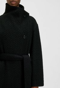 Czarny płaszcz o fakturowanej powierzchni z wysokim kołnierzem, dwoma guzikami i paskiem. Charakteryzuje się miękkim, puszystym materiałem oraz szerokimi rękawami.