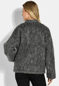 Vrouw met blond haar, gekleed in een losse, zachte donkergrijze trui en zwarte broek, met haar rug naar de camera gekeerd tegen een witte achtergrond.