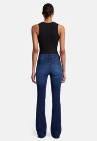 Jean évasé bleu marine à la texture lisse, avec une taille mi-haute et deux poches arrière. Porté avec un haut sans manches noir.