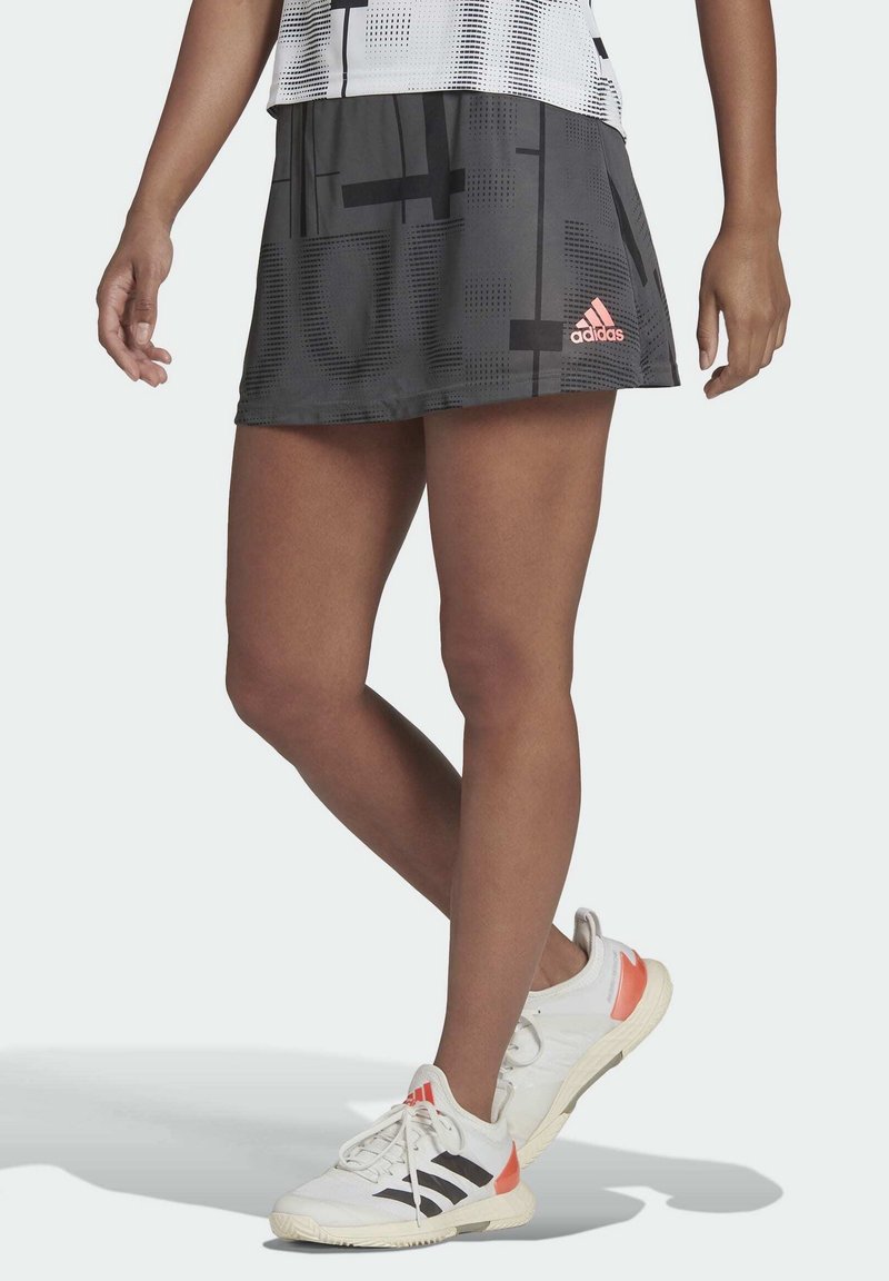 Svart sportkjol med geometriska mönster; har en rosa Adidas-logotyp. Bärs med vita skor, vilket visar en bekväm design och passform.