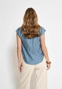 Top blu, a maniche corte con orlo arrotondato e leggermente stropicciato, abbinato a pantaloni a gamba larga di colore chiaro.