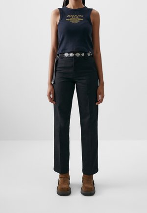 Personne portant un haut sans manches bleu marine Dickies avec logo doré, un pantalon noir à jambes droites, une ceinture à motifs et des bottines en daim marron, posant devant un fond blanc.