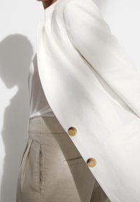 Osoba w beżowych, wysokich spodniach i białym żakiecie z jasnobrązowymi guzikami rzucająca cień na białym tle.