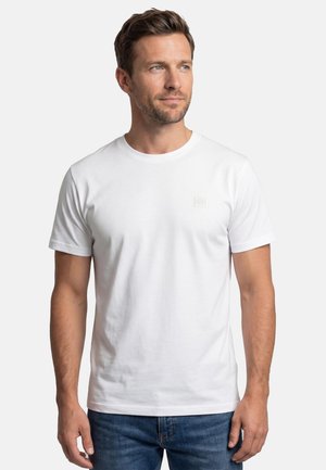 Mann mit kurzen Haaren, der ein schlichtes weißes T-Shirt mit kurzen Ärmeln und blaue Jeans trägt, leicht nach rechts blickend, stehend vor weißem Hintergrund.