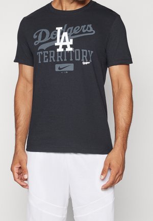 Mann trägt ein schwarzes Dodgers Territory T-Shirt mit weißem LA-Logo und Nike-Swoosh, kombiniert mit weißen Shorts.