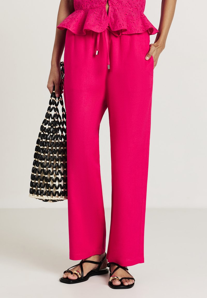 Fuchsia weit geschnittene Hose mit Kordelzugbund, kombiniert mit einer schwarzen geflochtenen Handtasche. Die Sandalen haben dünne schwarze Riemen und subtile metallische Details.