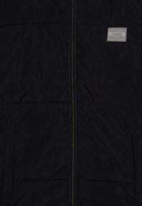 Veste matelassée noire avec fermeture éclair centrale et deux poches latérales, avec un petit patch rectangulaire sur la partie supérieure droite de la poitrine.