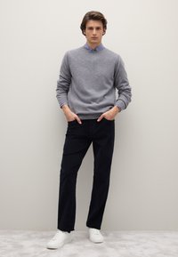 Maglione di lana grigio chiaro indossato sopra una camicia a righe blu e bianche, abbinato a pantaloni blu scuro e sneakers bianche. Design semplice e classico.