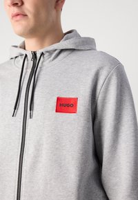 Šedá mikina s kapucí a předním zipem, na které je červená obdélníková nášivka označená "HUGO" a černé šňůrky. Měkčená textura látky.