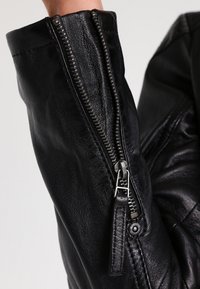 Black leather jacket sleeve featuring a prominent zipper detail, silver hardware, and textured finish, showcasing smooth and rugged characteristics.