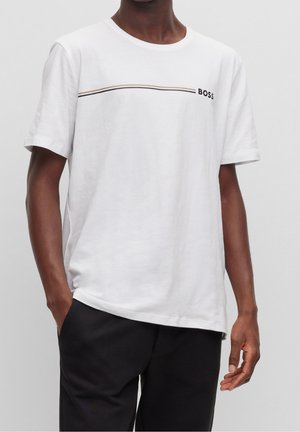 Mężczyzna w białej, krótkorękawowej koszulce z cienkimi poziomymi paskami i napisem "BOSS", w czarnych spodniach, stojący z jedną ręką w kieszeni.