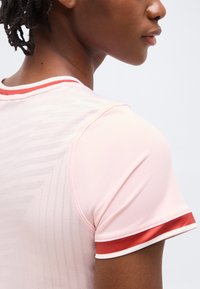 Približen posnetek osebe, ki nosi svetlo roza športno majico z rdečo in belo črto na rokavu ter teksturiranim zadnjim delom.