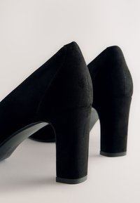 Coppia di scarpe con tacco alto in camoscio nero con punte arrotondate e tacchi a blocco robusti su sfondo chiaro.