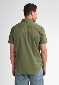 Camisa de manga corta de color verde oliva, fabricada en tejido ligero, con cuello abotonado, dobladillo curvado y detalles en el yugo trasero. Llevada con vaqueros azul claro.
