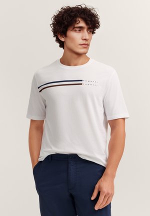 Mann mit lockigem Haar, der ein weißes kurzärmeliges Hemd mit marineblauen und braunen Streifen und marineblaue Hose trägt, steht mit den Händen in den Taschen und schaut nach rechts.