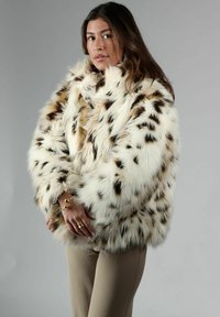 Faux fur-kappa i kräm med bruna fläckar, med en plyschig textur och oversized passform. Modellen bär beige byxor och guldsmycken.