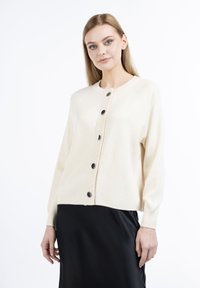 Cardigan crema realizzato in materiale morbido e leggero. Presenta polsini a coste, colletto rotondo e sei bottoni neri lucidi sul davanti.