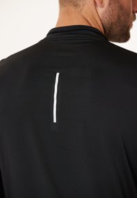 Sort atletisk skjorte med en glat tekstur. Har en slank hvid lodret streg på ryggen. Enkel design, crew neck stil.
