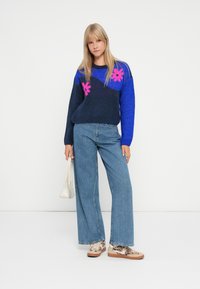 Knit sweater in blue with pink floral patterns, paired with loose-fitting denim jeans and textured sneakers. Model holding a white handbag.
