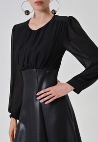 Zwarte jurk met doorzichtige, lange mouwen en een strakke bovenkant. De rok heeft een lichte glans, met een minimalistisch ontwerp en subtiele details.