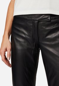 Zwarte leren broek met een gladde textuur, strak ontwerp en gestikte details op de tailleband, weergegeven met een hand die aan de zijkant rust.