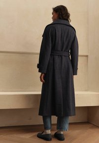 Long dark checkered trench coat with a belt, wide collar, and buttoned cuffs, paired with cropped denim pants and dark shoes.