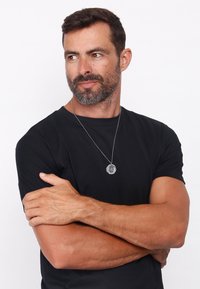 Sølv vedhængs halskæde på en kæde, båret over en sort t-shirt. Vedhænget har et cirkulært design med prægede detaljer.