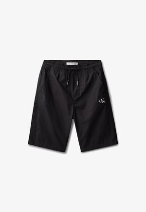 Niño con cabello rizado que lleva una chaqueta negra, camiseta blanca con el logo "Calvin Klein", pantalones cortos negros, calcetines blancos y zapatillas blancas, de pie con las manos en los bolsillos.