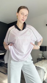 Ανοιχτό ροζ αθλητική φανέλα με μαύρο γιακά και λεπτομέρειες, featuring το κείμενο "ΑΣ ΓΙΝΕΙ" σε λευκό. Κοντά μανίκια, χαλαρή εφαρμογή, μαλακό ύφασμα.